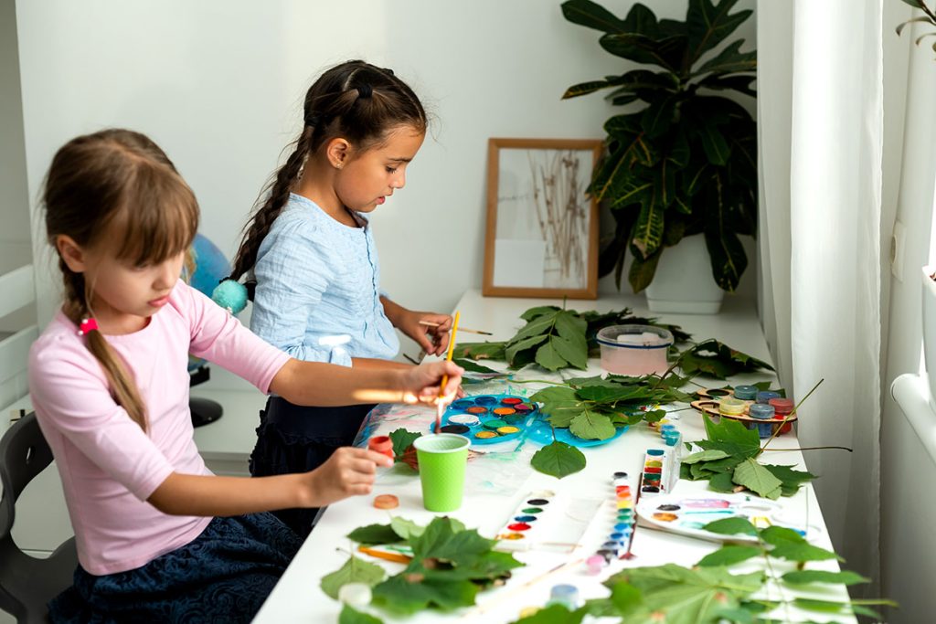 2 girls doing arts and craft projects with paints and leaves and different materials.